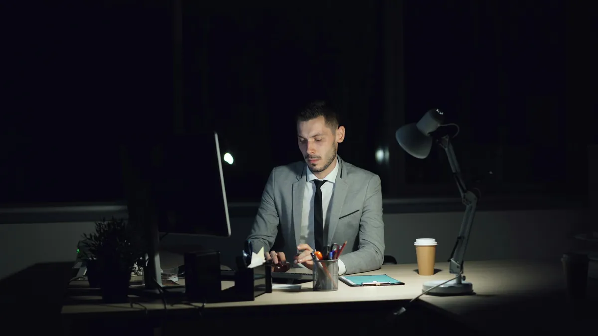 A banker in a suit working alone at a desk late at night under a desk lamp.