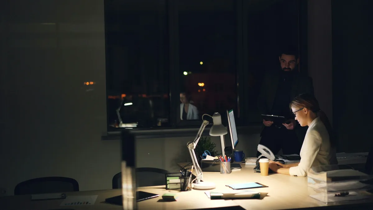 A dark office at night: one person at a desk under a lamp, a colleague standing behind.